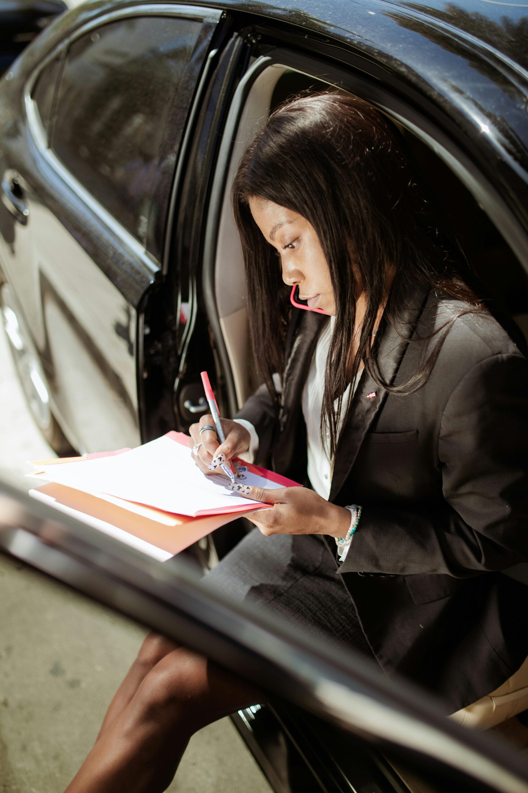 A professional woman in a black blazer sits in the open door of a car, writing on documents with a red pen.
